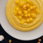 Photo of delicious vegan hummus on a white plate served with chickpeas and olive oil and surrounded by raw chickpeas, garlic cloves and lemon.