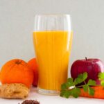 A photo of a glass with an orange smoothie surrounded by oranges, apple, carrot, celery, ginger and flax seeds. A vegan recipe from All About Vegans.