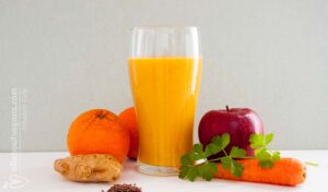 A photo of a glass with an orange smoothie surrounded by oranges, apple, carrot, celery, ginger and flax seeds. A vegan recipe from All About Vegans.