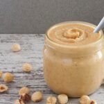 Photo of homemade hazelnut butter in a jar, surrounded by hazelnuts. A vegan and vegetarian recipe from All About Vegans.