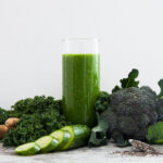 Photo of green smoothie in a transparent glass surrounded by broccoli, kale, ginger, cucumber and chia seeds.