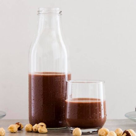 Photo of a glass bottle with homemade chocolate hazelnut milk, with a background of chocolate truffles that we make with whatever is left over from the hazelnuts. In the foreground, low, there are also raw hazelnuts. A vegan and homophagic recipe from All About Vegans com.